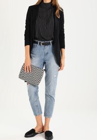 Blazer negro de manga larga sobre un suéter de cuello alto a rayas negras, combinado con jeans desgastados azul claro y mules planos negros. Sosteniendo un bolso de mano con diseño en zigzag.