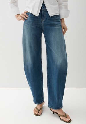 Person trägt lockere blaue Jeans, weißes langärmeliges Hemd und braune Sandalen mit niedrigem Absatz vor einem schlichten weißen Hintergrund.