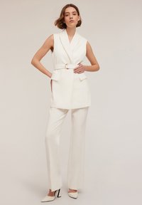 Sacou alb fără mâneci, cu curea, asortat cu pantaloni largi. Materialul este neted, cu un design structural și buzunare simple.