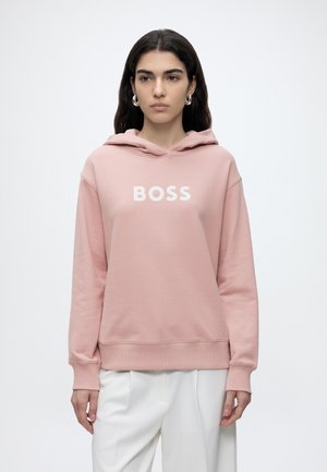 Jeune femme aux longs cheveux foncés portant un sweat à capuche rose avec le mot BOSS et un pantalon blanc, debout devant un fond blanc uni.