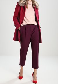 Punainen villatakki, jossa on karvakaulus, vaaleanpunainen poolopaita ja maroon-väriset laskostetut housut. Yhdistetty punaisiin korkokenkiin. Suorat pinnat, istuva leikkaus.