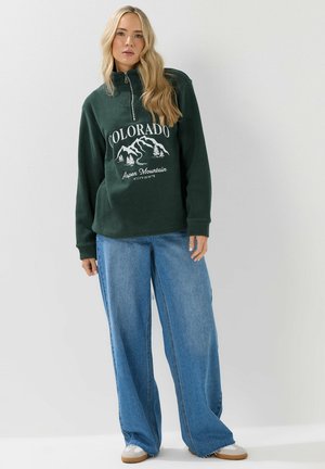 Felpa verde in pile con zip a metà davanti e grafica "COLORADO"; abbinata a jeans in denim blu a gamba larga; visibile calzature casual.