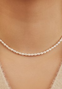 Collier de perles avec de petites perles ovales, brillantes et blanches, régulièrement enfilées ensemble, posées sur un fond de teint neutre.