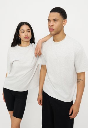 Une femme pose son bras sur l'épaule d'un homme ; tous deux portent des t-shirts gris clair et des bas noirs, sur un fond clair uni.