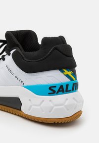 Zapatilla deportiva en blanco y negro con acentos azules. Presenta una tela texturizada, un talón acolchado y la marca "SALM" en el lateral.