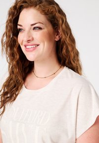 T-shirt beige clair avec des manches retroussées, mettant en avant un imprimé texturé. Le modèle porte un collier en or délicat agrémenté de petites accents ronds.