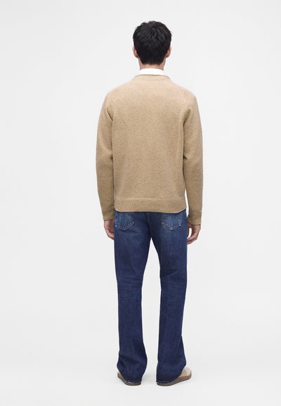 Homme debout de dos portant un pull beige, un jean bleu et des chaussures beige sur un fond blanc uni.