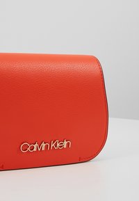 Pochette en cuir rouge avec logo doré "Calvin Klein". Présente une texture lisse et une forme structurée, avec des bords arrondis et une fermeture à rabat.