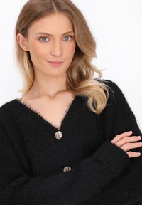 Cardigan nero lavorato a maglia con scollo a V, caratterizzato da un motivo testurizzato e un dettaglio con bottone argentato sul petto. Maniche lunghe coprono i polsi.