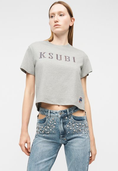 Γυναίκα που φοράει γκρι cropped t-shirt Ksubi και μπλε τζιν με κομμένες ζώνες στη μέση και κρυστάλλινες διακοσμήσεις στις μπροστινές τσέπες.