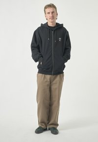 Schwarze Zip-Hoodie mit Kängurutaschen und Kordelzügen, kombiniert mit beigen Hosen und grünen Turnschuhen. Verfügt über ein kleines Grafiklogo auf der Brust.