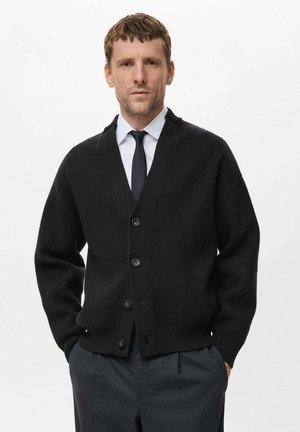 Cardigan noir en tricot ; présente un col en V, des manches longues et six boutons sur le devant. Porté par-dessus une chemise blanche et une cravate.