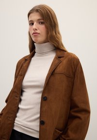 Brown suede blazer with notched lapels, single button closure, and chest pocket, layered over a light grey turtleneck top.