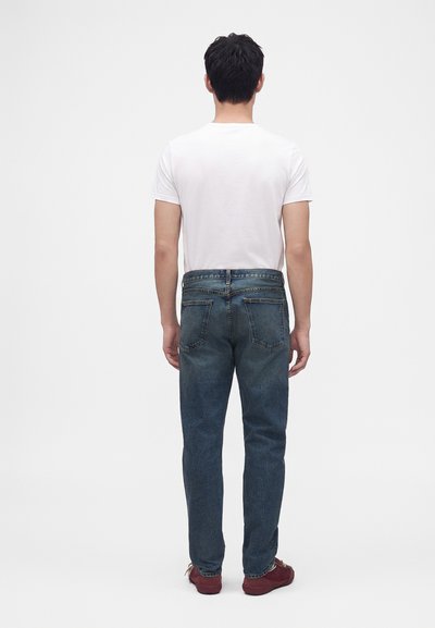 Homme debout de dos portant un t-shirt blanc uni, un jean bleu et des chaussures rouges sur un fond blanc uni.