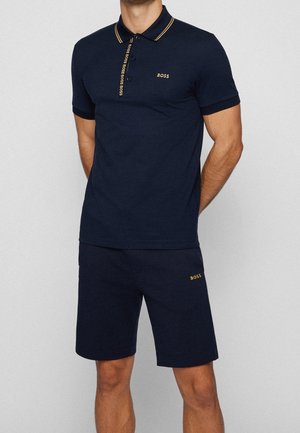 Mężczyzna w granatowej koszulce polo z krótkim rękawem i dopasowanych szortach z złotymi logo "BOSS" oraz paskowanym kołnierzykiem na jednolitym tle.