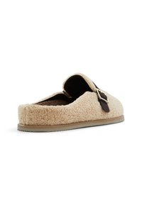 Beige slipper met een pluche textuur, open achterkant ontwerp, bruine gespsluiting en rubberen zool, met een zachte, donzige bovenkant.