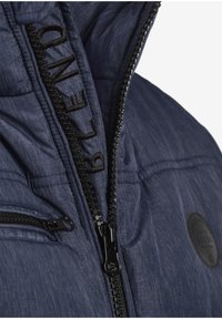 Marineblå puffjakke med teksturert stoff, svart glidelås, brodert logo og sidelomme med glidelåslukking.