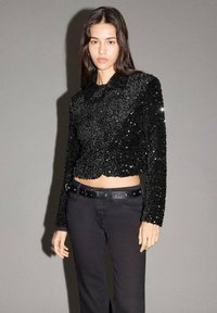 Giacca corta nera con paillettes e maniche lunghe, caratterizzata da un colletto e un'apertura con bottoni. Abbinata a pantaloni neri e una cintura nera.