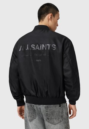 Man wearing a black bomber jacket with "ALLSAINTS" and "LONDON" text printed on the back, paired with faded grey jeans.