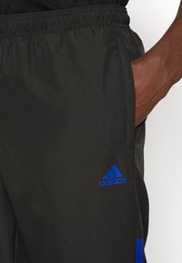 Pantalones deportivos negros con cintura elástica, que presentan el logo azul de Adidas y acentos azules en los lados de la pierna. Material liso y ligero.