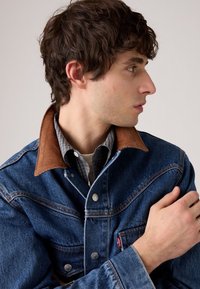 Joven con cabello rizado oscuro que lleva una chaqueta de mezclilla azul con cuello marrón y una camisa de cuadros, mirando hacia su derecha.