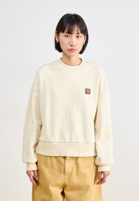 Sweatshirt couleur crème avec des poignets et un ourlet côtelés, arborant une petite étiquette en cuir de la marque sur la poitrine ; coupe décontractée et texture douce.