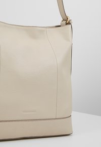 Beige läderhandväska med en texturerad yta, med en platt botten, subtila sömmar och ett guldfärgat metallfäste på axelremmen.