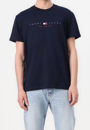 Mand iført en marineblå Tommy Jeans T-shirt med logo og lyseblå jeans, stående foran en ensfarvet hvid baggrund.