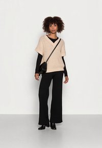 Camisola de malha bege oversize de mangas curtas, usada sobre uma camisa preta de mangas compridas, combinada com calças pretas de perna larga e botas de salto.