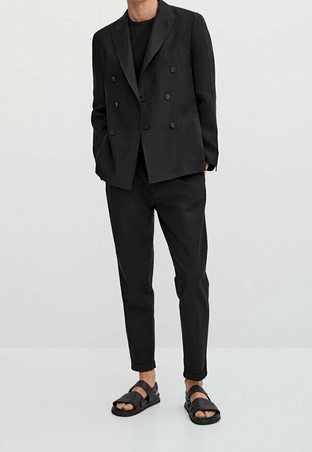 Blazer noir à double boutonnage avec un haut en tricot texturé, associé à un pantalon noir ajusté et des sandales plates noires, créant un look monochromatique.
