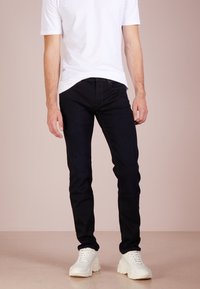 Jeans in denim nero con una vestibilità slim, abbinati a una t-shirt bianca a maniche corte e sneakers chunky bianche su uno sfondo neutro.