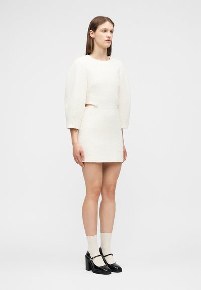 Γυναίκα στέκεται φορώντας λευκό μίνι φόρεμα με υφή και φουσκωτά μανίκια, με ανοίγματα στα πλάγια, κρεμ κάλτσες και μαύρες γόβες Mary Jane.