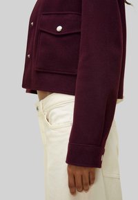 Burgundy cropped jacket with two front pockets, silver snap buttons, and a smooth texture, paired with cream-colored denim pants.
