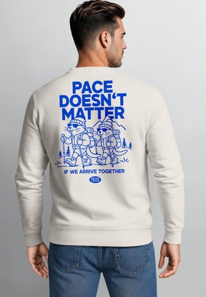 Man wearing a white sweatshirt with blue text and hiking otter cartoons reading "Pace doesn't matter if we arrive together."