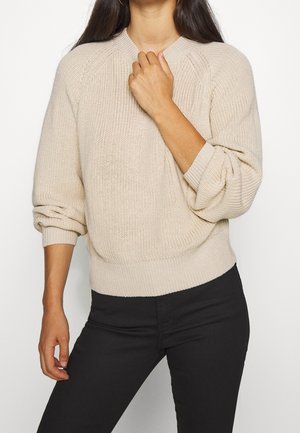 Kvinde iført en beige strikket sweater med opkørte ærmer og sorte bukser, med den ene hånd, der rører sweateren nær halsen.