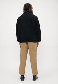 Mand med dreads iført sort fleecejakke, beige cargo bukser og sorte sko, står med ryggen til på hvid baggrund.