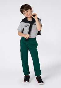 Jeune garçon portant un t-shirt gris Champion, un pantalon cargo vert, des baskets noires, avec un sweat-shirt noir drapé sur les épaules, se tenant sur un fond blanc.
