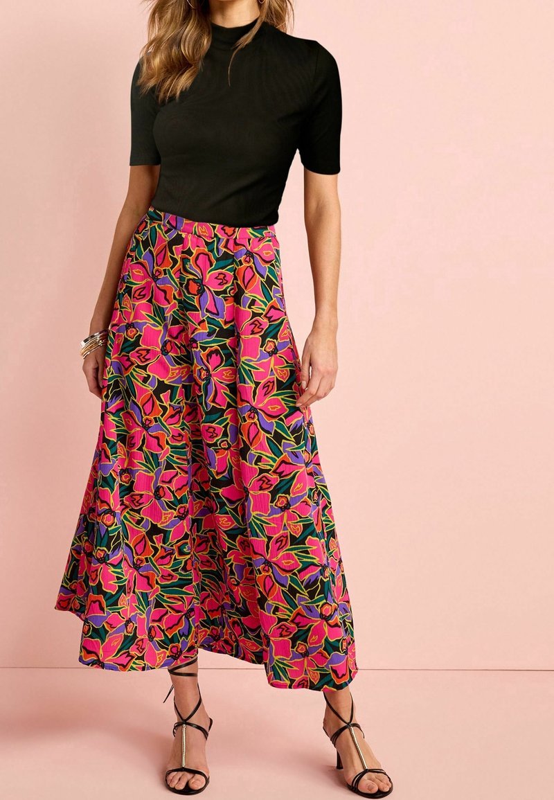 Femme portant un haut noir à manches courtes et un pantalon taille haute à jambes larges avec un motif floral rose vif, vert et violet, debout sur un sol rose.