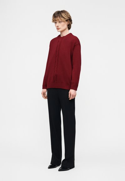 Άτομο με κοντά μαλλιά που φοράει μπορντό πουλόβερ με πλεκτό σχέδιο, μαύρο παντελόνι και μαύρα παπούτσια, όρθιο μπροστά σε λευκό φόντο.