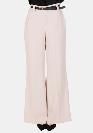 Donna che indossa pantaloni beige a vita alta e gamba larga con cintura nera e tacchi neri a punta, in piedi su sfondo bianco.