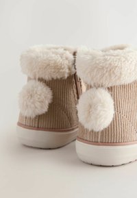 Beżowe buty z kordurowej tkaniny z puszystym kremowym mankietem, dwiema dużymi pomponami po bokach i białą gumową podeszwą. Teksturowana powierzchnia materiału.