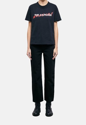 Person wearing black pants, black boots, and a black t-shirt with red and white overlapping text across the chest.