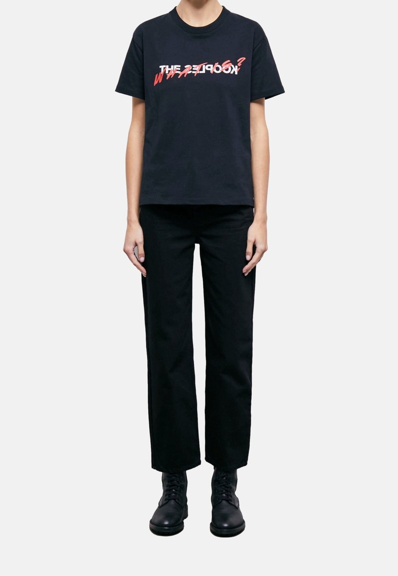 Osoba ubrana w czarne spodnie, czarne buty i czarną koszulkę z czerwonym i białym nakładającym się napisem na piersi.