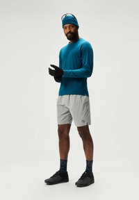 Turkooise langemouw shirt, lichtgrijze shorts, zwarte handschoenen en donkere sneakers; gestructureerde gebreide beanie en gestreepte sokken met atleetontwerpdetails.