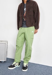 Brun fleecejacka med dragkedjefickor, gröna cargo-byxor med sidofickor, sneakers i svart och neongrön accent, stående på blå matta.