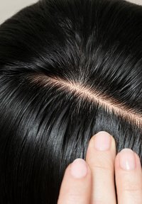 Des cheveux noirs raides sont montrés de près, avec une raie visible. Une main, avec une peau claire et des ongles courts, touche doucement les cheveux.
