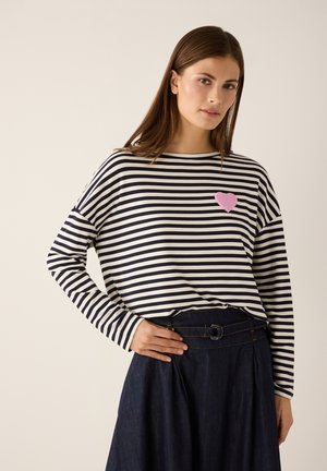 Frau trägt ein langärmeliges Oberteil mit schwarz-weißen Streifen und einem pinkfarbenen Herzfleck sowie einen dunkelblauen, hoch taillierten Rock mit Gürteldetail.