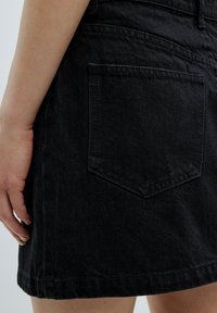 Schwarzer Jeansrock mit einer hinteren Tasche und spitzem Klappverschluss, sichtbaren Nähten und geradem Saum. Hand greift den Seitenteil des Rocks.