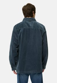 Mand iført en løs, mørkeblå corduroyjakke og blå jeans, vendt væk, der viser bagsiden af jakken og overkroppen.