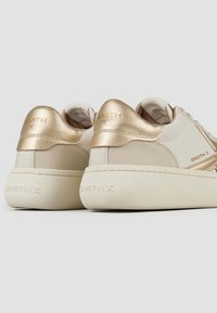 Sneaker beige e oro con finitura in pelle liscia, dotate di un tallone imbottito dorato e dettagli testurizzati. Marchio visibile sul tallone.